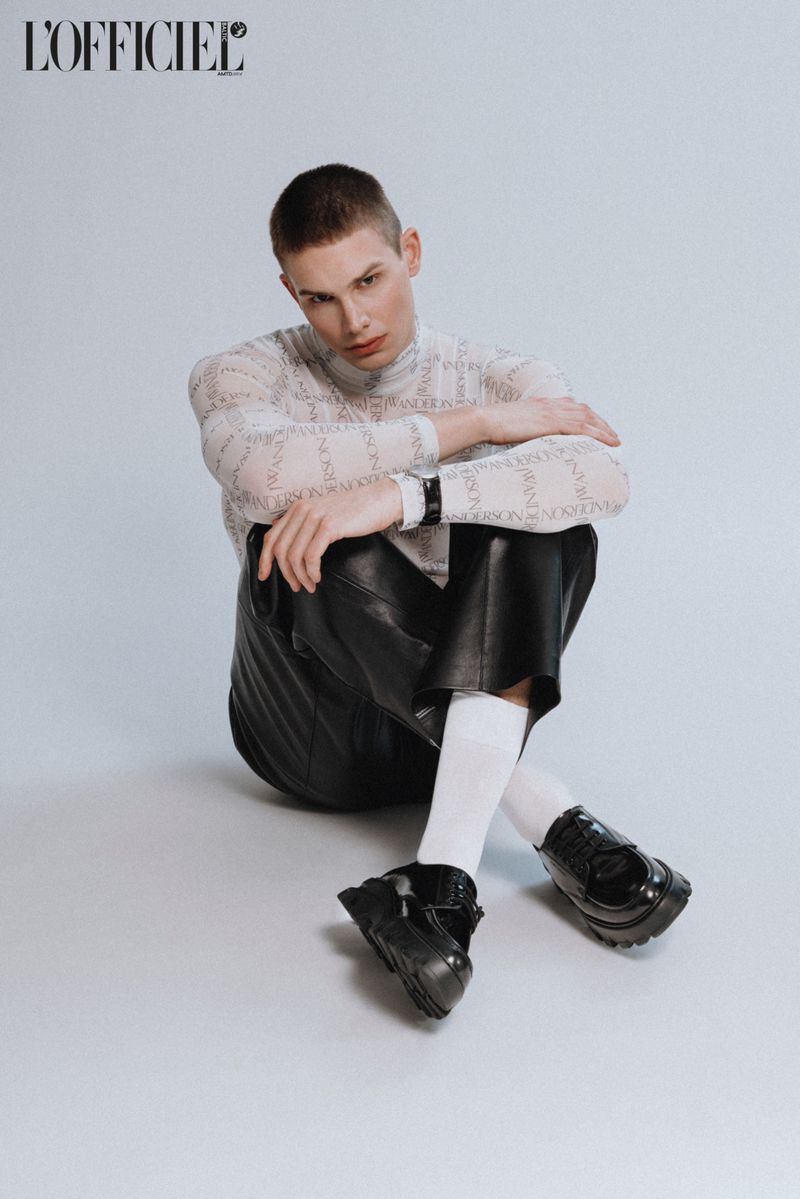 A person with short hair sits on a white surface wearing a patterned long-sleeve shirt, black leather shorts, white socks, and black shoes.