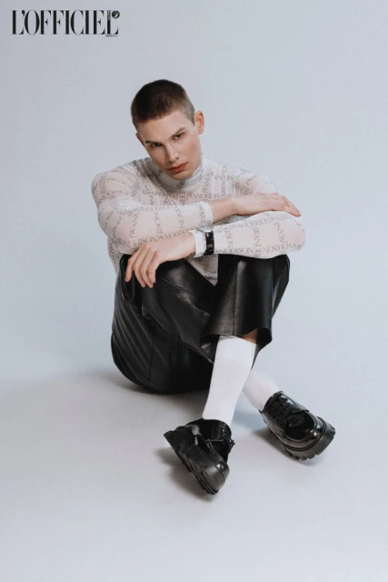 A person with short hair sits on a white surface wearing a patterned long-sleeve shirt, black leather shorts, white socks, and black shoes.