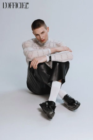 A person with short hair sits on a white surface wearing a patterned long-sleeve shirt, black leather shorts, white socks, and black shoes.