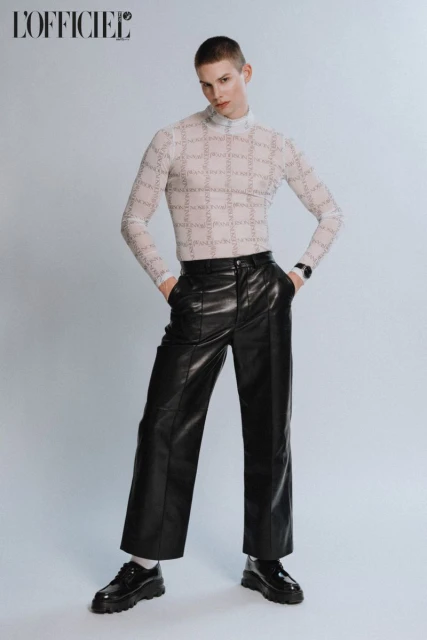 A fair-skinned male model poses in a studio, wearing a patterned sheer top, black leather pants, and black platform shoes for a fashion editorial.
