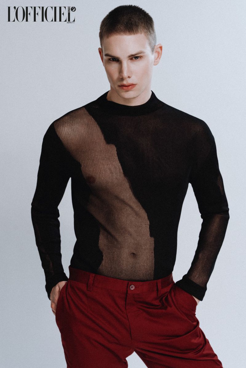 Fashion shot of a slender man in a sheer black top and dark red pants against a clean white backdrop, minimalist studio style.