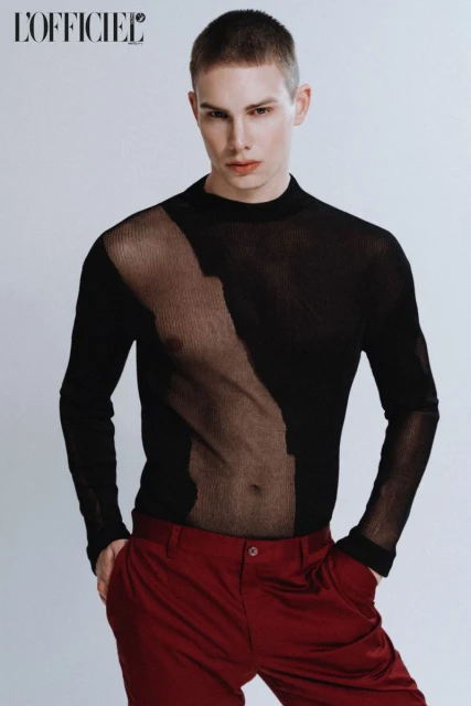 Fashion shot of a slender man in a sheer black top and dark red pants against a clean white backdrop, minimalist studio style.