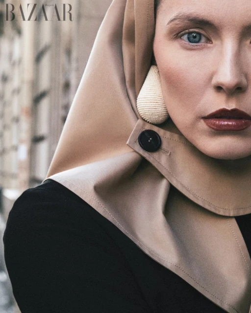 Close-up of a woman with blue eyes wearing a hooded garment with a button, complemented by large earrings and striking red lipstick.