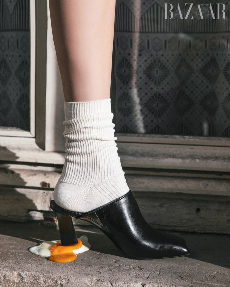 Fashion-forward editorial shot features a black mule, white socks, and cracked egg near a patterned wall and 'BAZAAR' logo.
