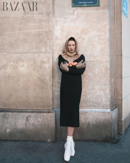 A model in a black dress and beige head covering stands holding oranges, posing against a textured wall on a city street.