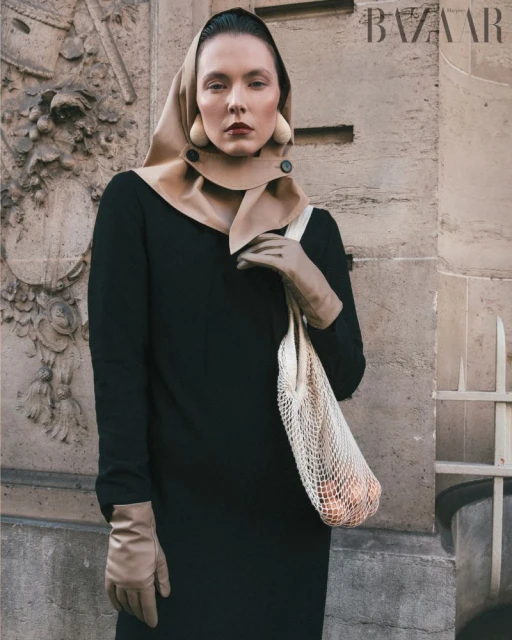 A woman in a black dress and headscarf stands next to a textured wall holding a mesh bag in a medium shot with muted colors.