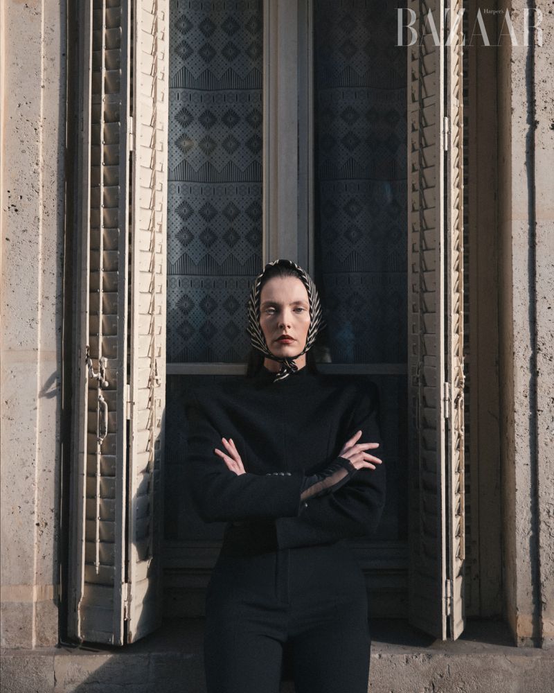 Fashion portrait of a woman wearing a headscarf and a black suit, posing near a window with shutters, bathed in soft, diffused light.