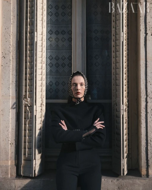 Fashion portrait of a woman wearing a headscarf and a black suit, posing near a window with shutters, bathed in soft, diffused light.