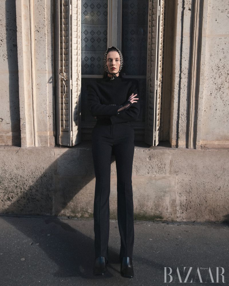 Fashion shot of a model in a black pantsuit and headscarf posing in front of a tall window in an urban setting with natural light.