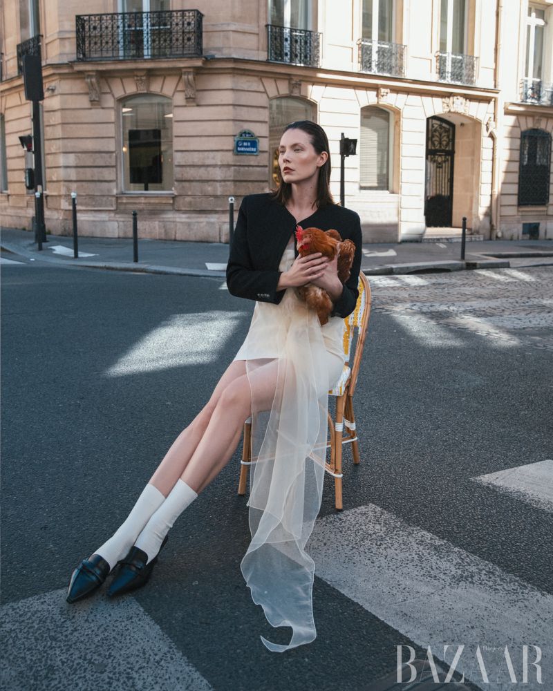 Fashion shot of a woman sitting on a chair on a pedestrian crossing in Paris, holding a live chicken, wearing a black jacket and dress.