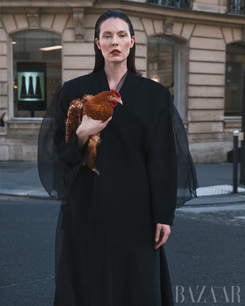 A fashion portrait captures a woman in a black dress holding a chicken on a city street during the day. She is posing for the camera.