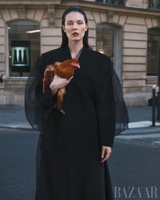A fashion portrait captures a woman in a black dress holding a chicken on a city street during the day. She is posing for the camera.