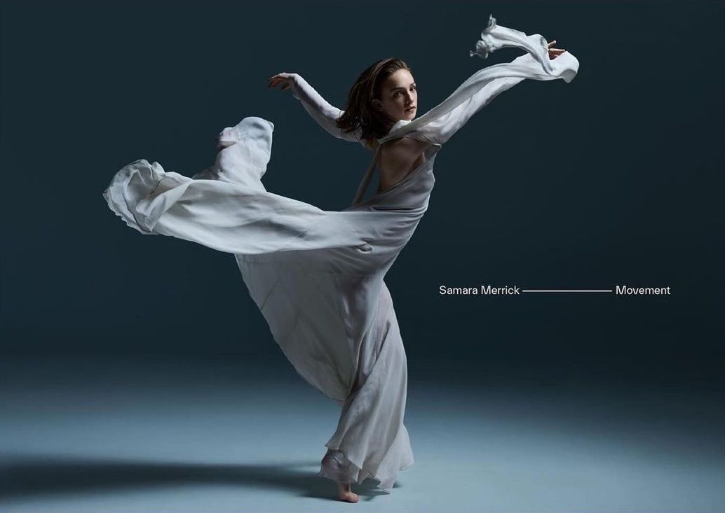 Female dancer in flowing white dress strikes dynamic pose in studio against a gray background, embodying movement and grace in a captivating image.