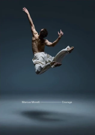 Athletic man in mid-air performing dance routine against gray backdrop, wearing white pants, arms extended, lit from above.