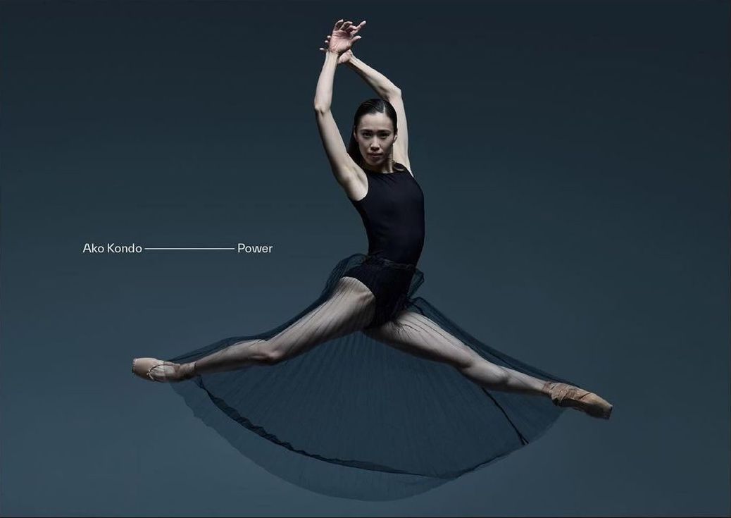Female ballet dancer in mid-air leap wearing a black leotard and sheer skirt against a grey background, in a dance or fitness pose.