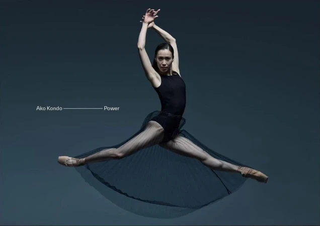 Female ballet dancer in mid-air leap wearing a black leotard and sheer skirt against a grey background, in a dance or fitness pose.