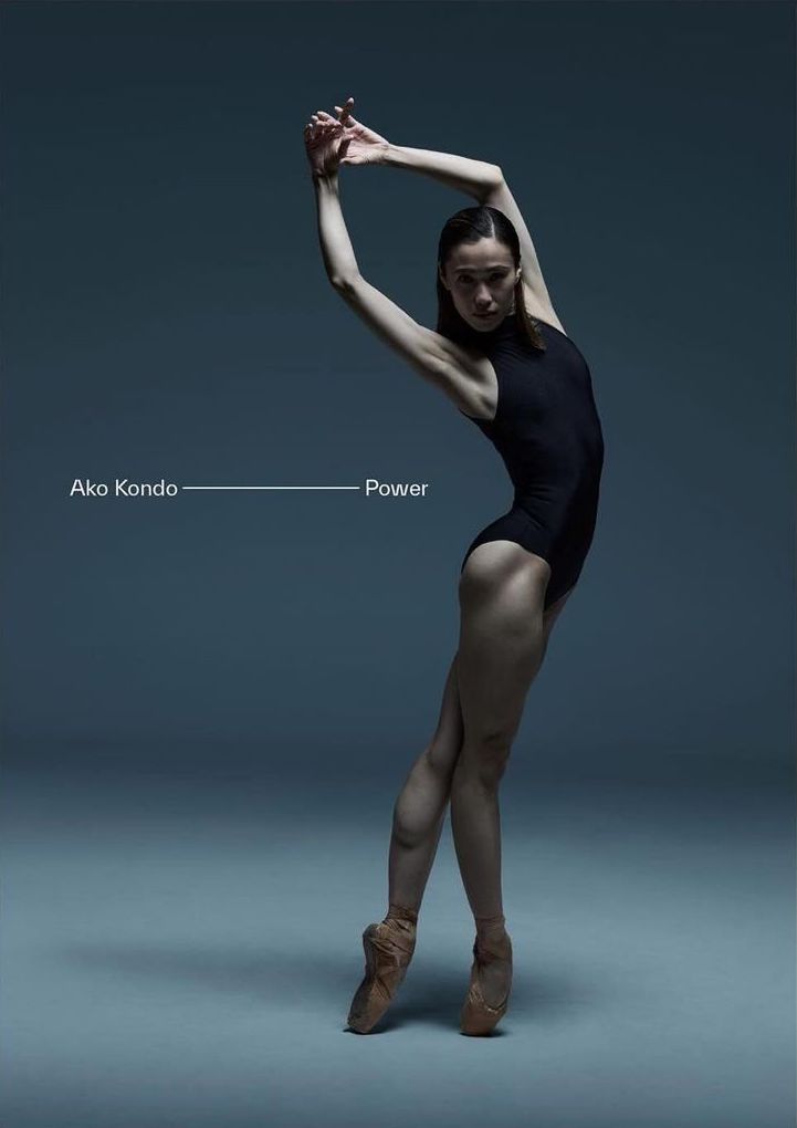 Female dancer in a black leotard and pointe shoes arches backward with her arms raised in a studio with a grey background and dramatic lighting.