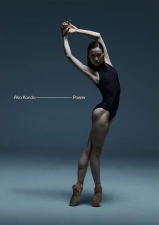 Female dancer in a black leotard and pointe shoes arches backward with her arms raised in a studio with a grey background and dramatic lighting.