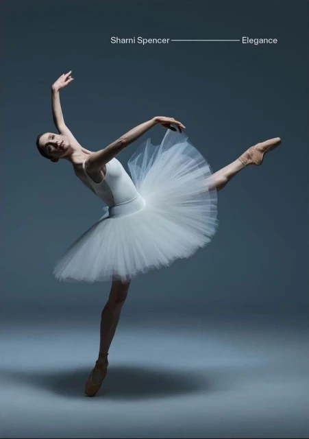 Female ballet dancer in a white tutu and pointe shoes poses gracefully against a gray background in a dance studio.