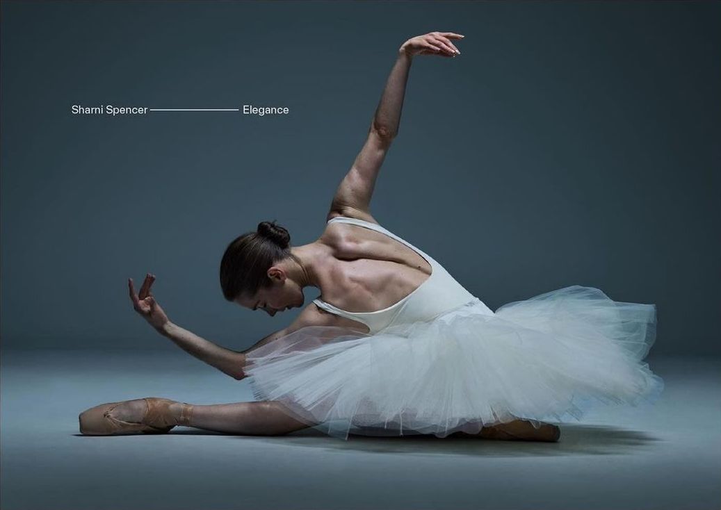 Ballet dancer in white tutu performing a split on a gray floor in a photo studio with professional lighting and dramatic posing.
