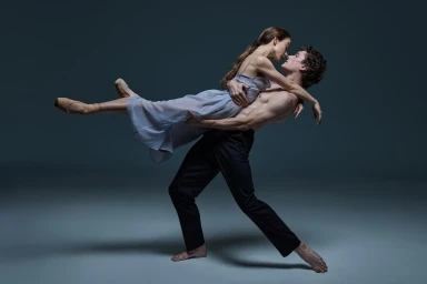 Two ballet dancers performing a dramatic lift against a muted dark studio backdrop, wearing light blue dress and dark trousers.
