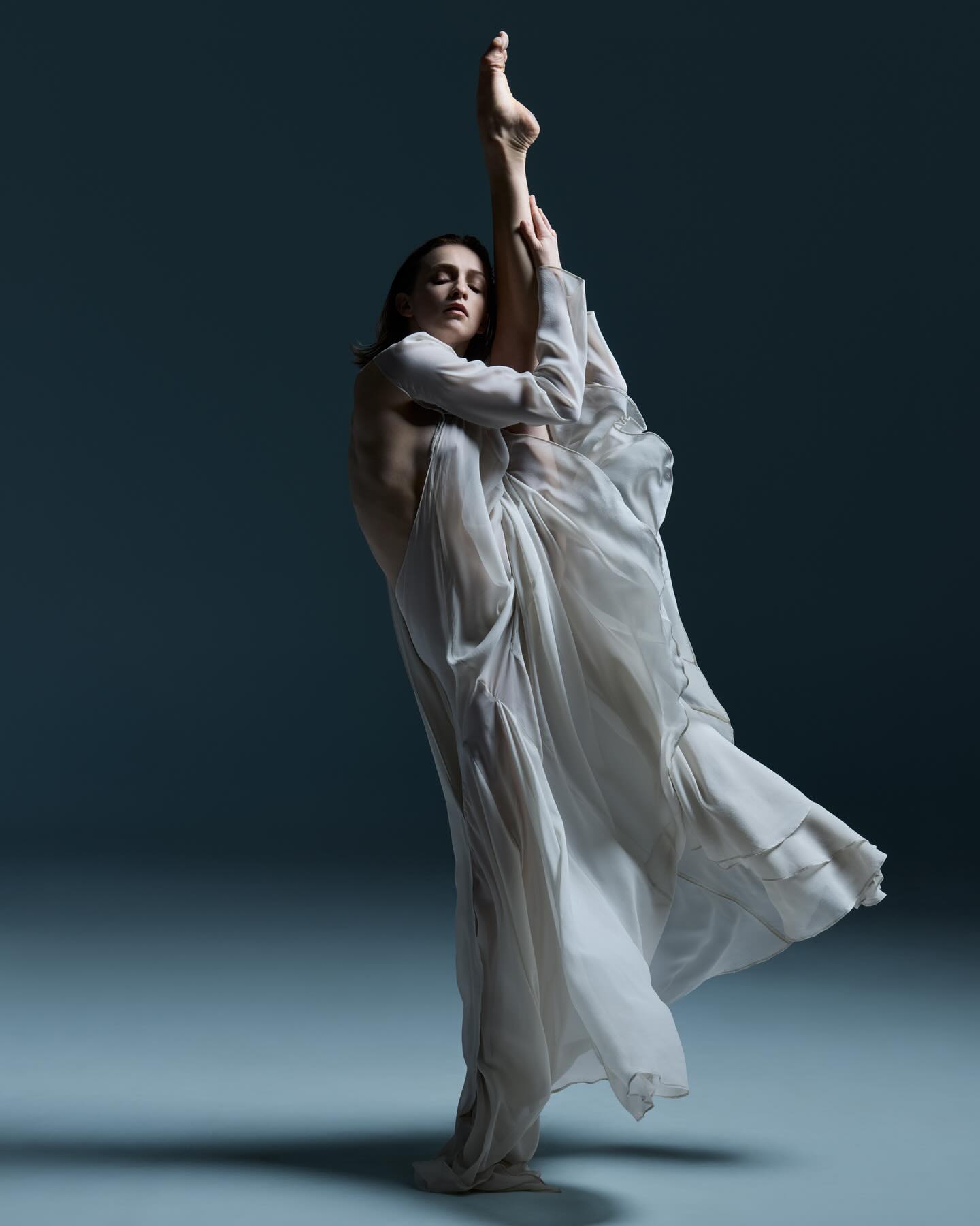 Adult female dancer in flowing white dress in a leg-up pose against a neutral background in a studio environment.