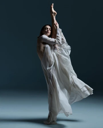 Adult female dancer in flowing white dress in a leg-up pose against a neutral background in a studio environment.