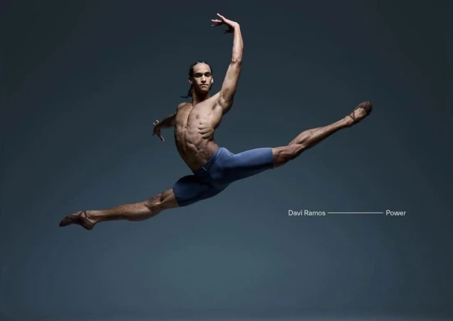 Male ballet dancer in mid-air leap against a dark grey background, wearing blue shorts and ballet shoes, muscular physique, artistic pose.