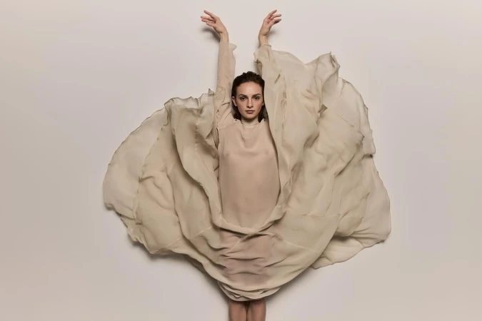 Woman in flowing beige fabric poses in a photo studio. The fabric surrounds her and extends above her head as she looks at the camera.