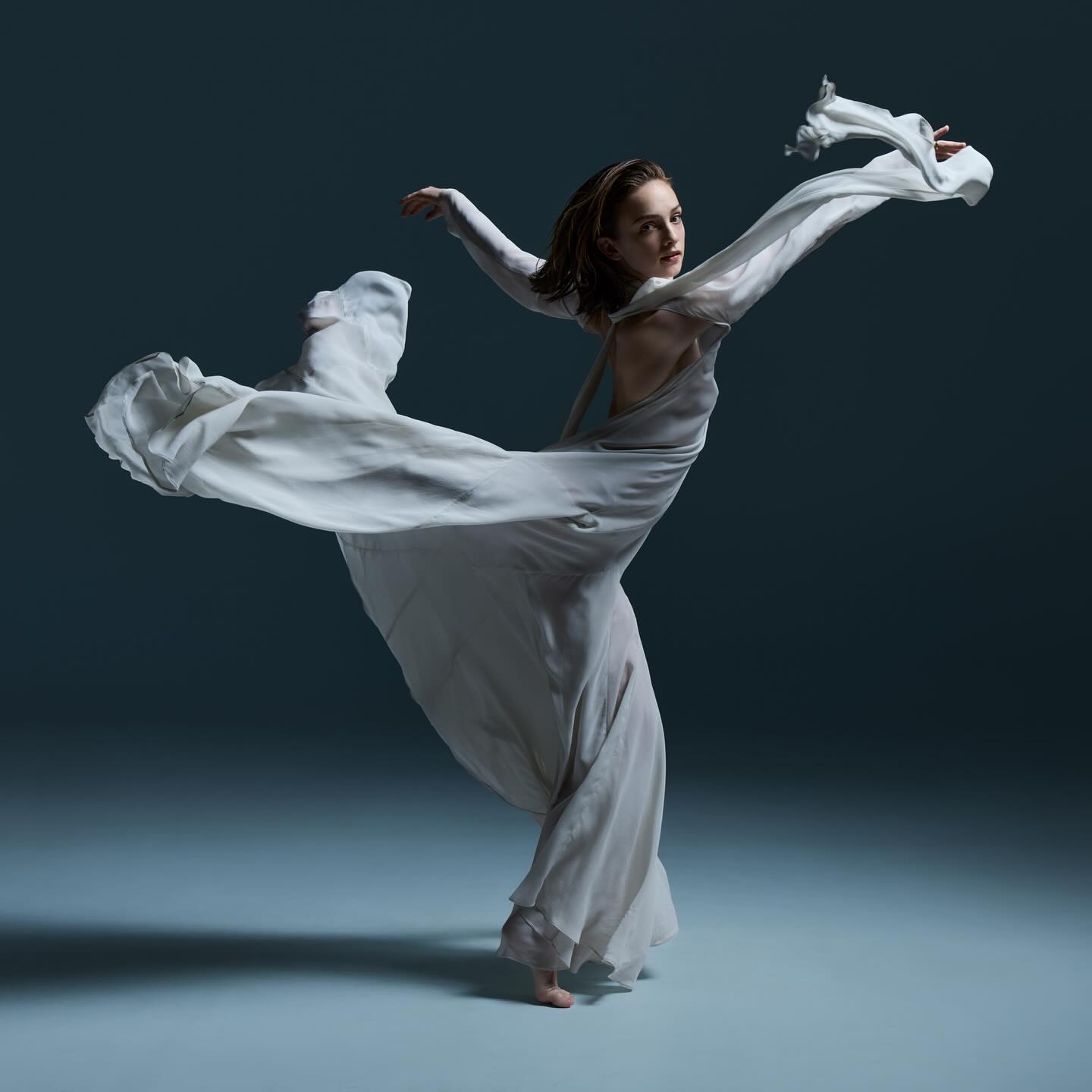 Woman in flowing white dress strikes a dance pose in a dimly lit studio against a dark blue backdrop, highlighting movement and grace.