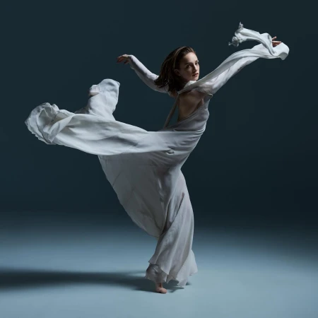 Woman in flowing white dress strikes a dance pose in a dimly lit studio against a dark blue backdrop, highlighting movement and grace.