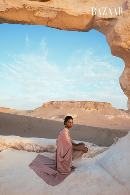 Woman in flowing pink fabric sits on a rock formation in a desert landscape under a stone arch, featuring Bazaar logo.