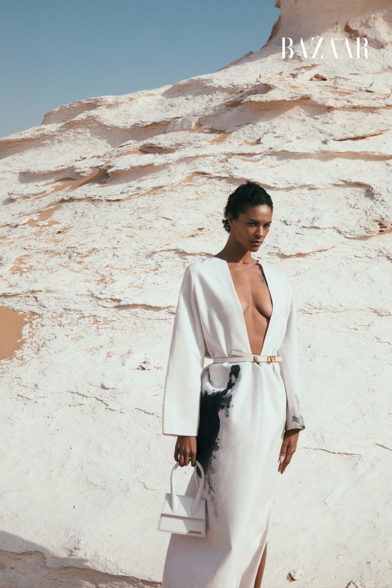 An adult female in a long white dress stands in front of a rock formation in a desert, holding a small white handbag in the sunlight.