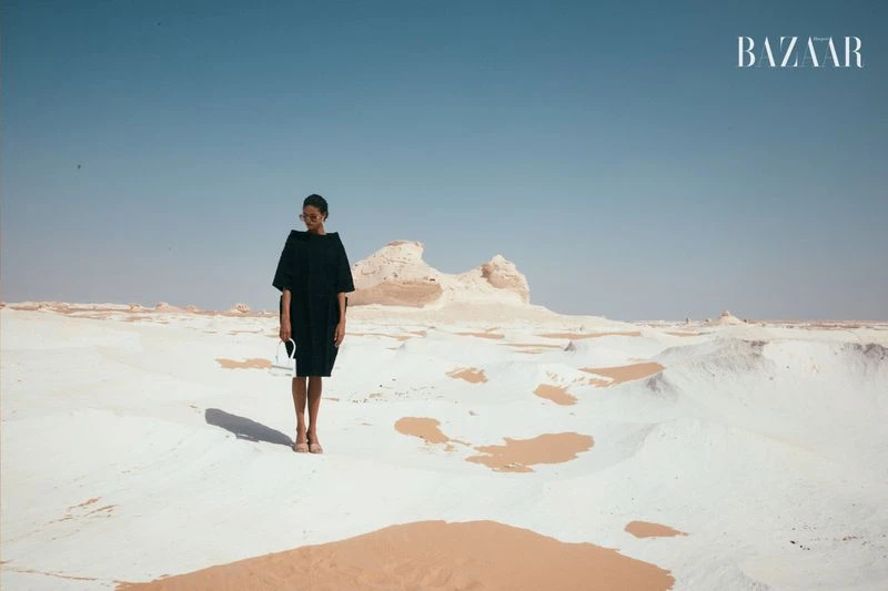 Fashion shot of a woman in a black dress and sunglasses standing in a desert landscape with white rock formations, under a clear blue sky.
