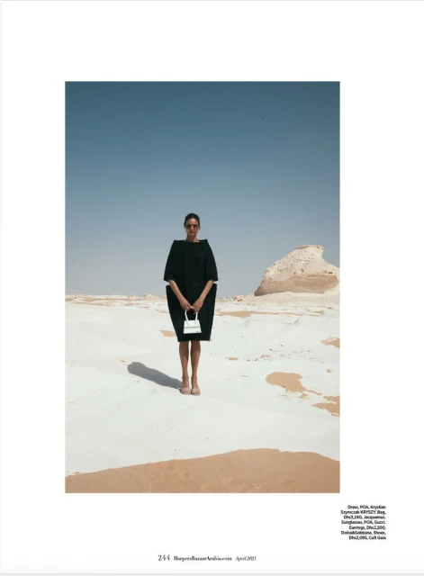 Fashion shot of a woman in a dark dress holding a white purse, standing in a desert landscape under a clear blue sky.
