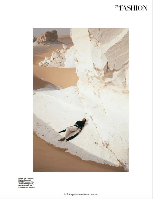 Fashion editorial shot of a woman lying on white rocks in the desert, wearing a dark top and light-colored pants, under natural sunlight.