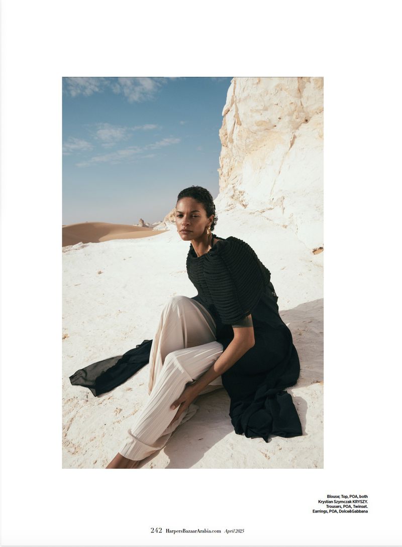 Woman poses in sand wearing a black top and tan pants with white rock formations and a tan desert backdrop under a bright blue sky.