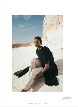 Woman poses in sand wearing a black top and tan pants with white rock formations and a tan desert backdrop under a bright blue sky.
