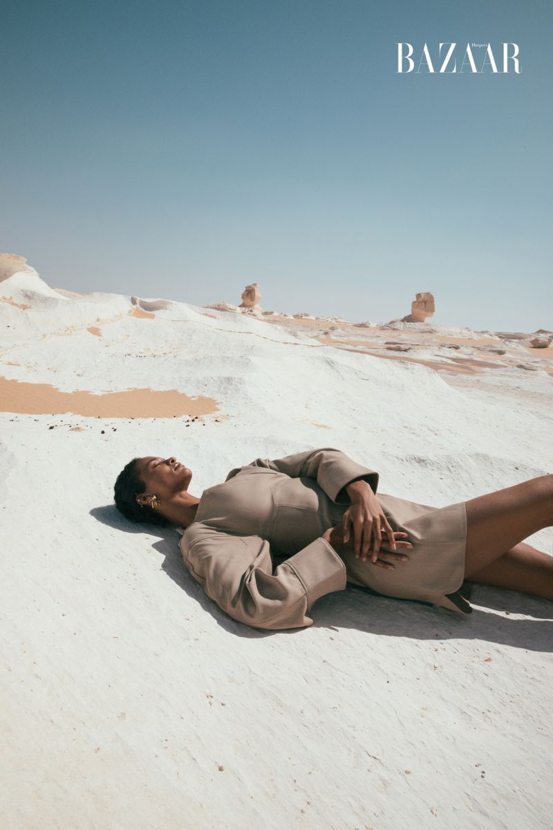 Woman in tan dress lies on white sand dunes in desert under a clear blue sky. Fashion editorial photo in a minimalist landscape.