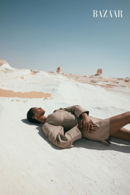 Woman in tan dress lies on white sand dunes in desert under a clear blue sky. Fashion editorial photo in a minimalist landscape.
