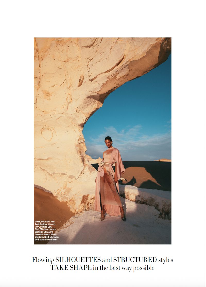 Fashion editorial shoot: woman in a long peach-colored dress stands beneath a rock archway with a view of blue sky and land.