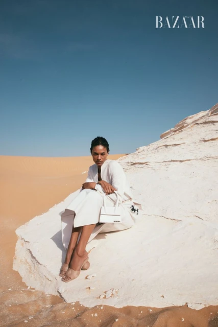 A woman in white sitting on a rock formation in a desert landscape wearing a matching bag and sandals, under a clear blue sky. 