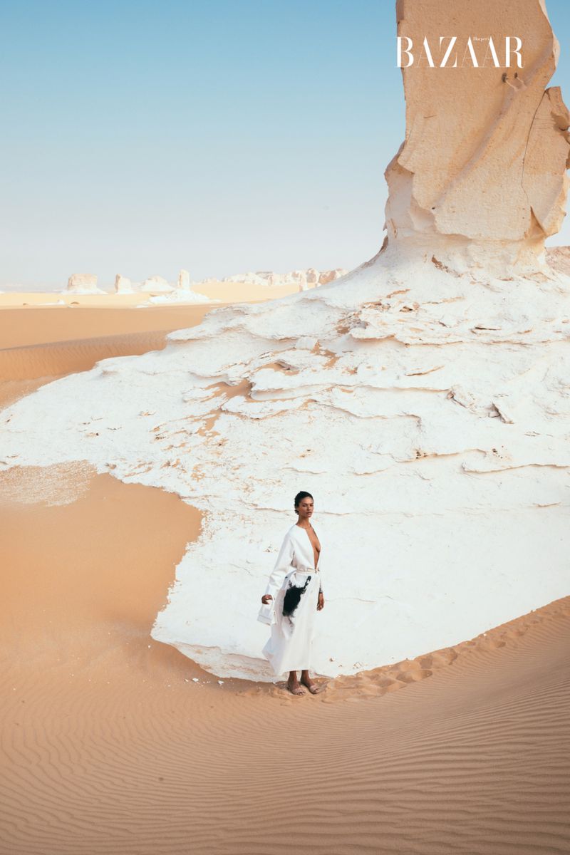 Fashion shot of a woman in a white robe in a sandy desert environment with white rock formations and a clear sky; Bazaar logo visible.