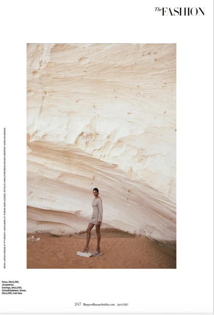 Fashion editorial shot: adult female model posing in a sandy outdoor setting wearing a neutral-toned dress in front of a layered rock formation.