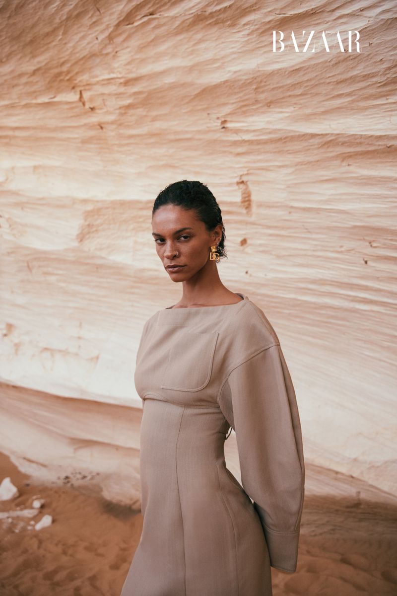 A model poses in a light brown dress against a textured rock formation for a fashion editorial, exuding understated elegance and earthy tones.
