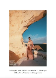 Model wearing a long pink dress standing beneath a natural rock arch formation with a clear blue sky and desert landscape background.