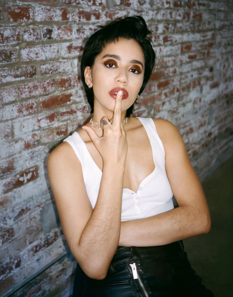 Portrait of a young woman with short dark hair posing indoors against a textured brick wall, wearing a white top and black skirt.