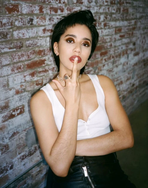Portrait of a young woman with short dark hair posing indoors against a textured brick wall, wearing a white top and black skirt.