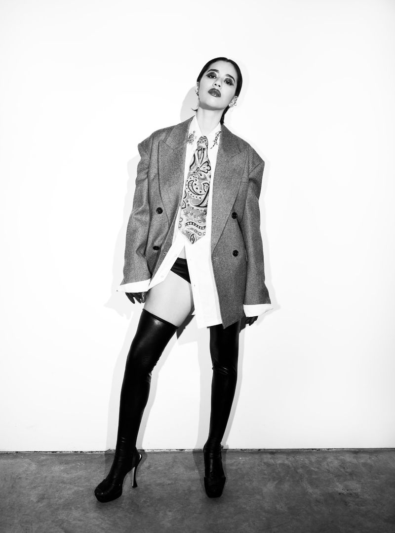 A full shot of a fashionable woman in a gray blazer, white shirt, tie, thigh high boots, and black underwear against a white background.