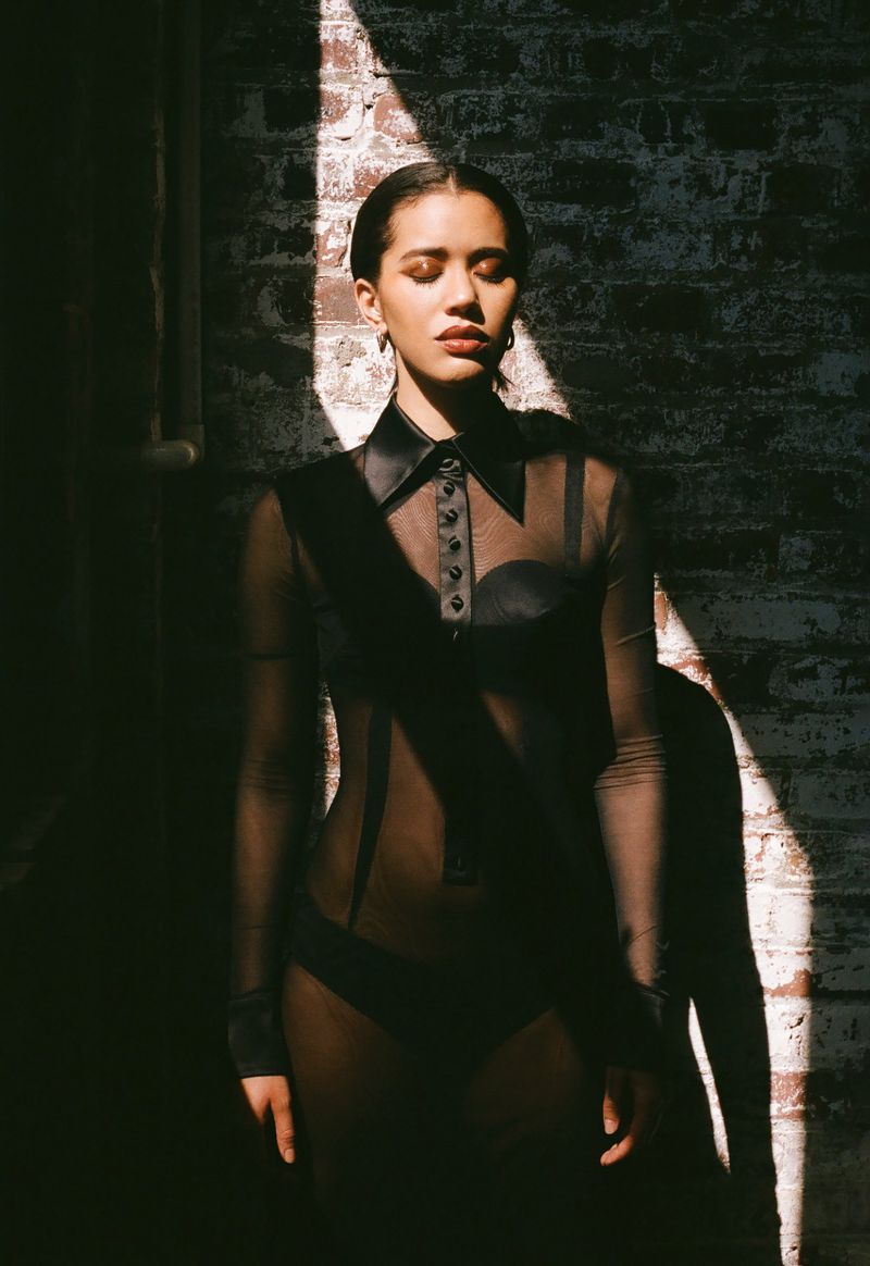 A young woman with her eyes closed stands in a beam of light against a brick wall. She's wearing a dark, sheer shirt with a collar.